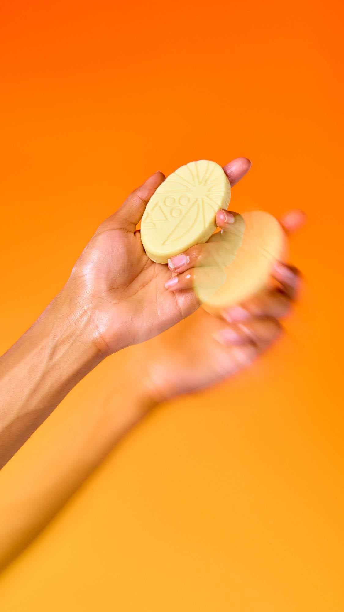 An oval, patterned cream Lush massage bar is held in a person's open hand against a vibrant orange background. A second hand is blurred in motion nearby, creating a dynamic effect.