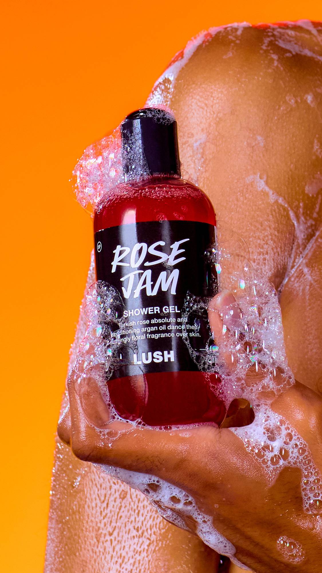 A hand holds a bottle of vibrant red Lush Rose Jam shower gel, covered in frothy, iridescent soap bubbles. The product is held against a wet, lathered shoulder and a vivid, solid orange background.