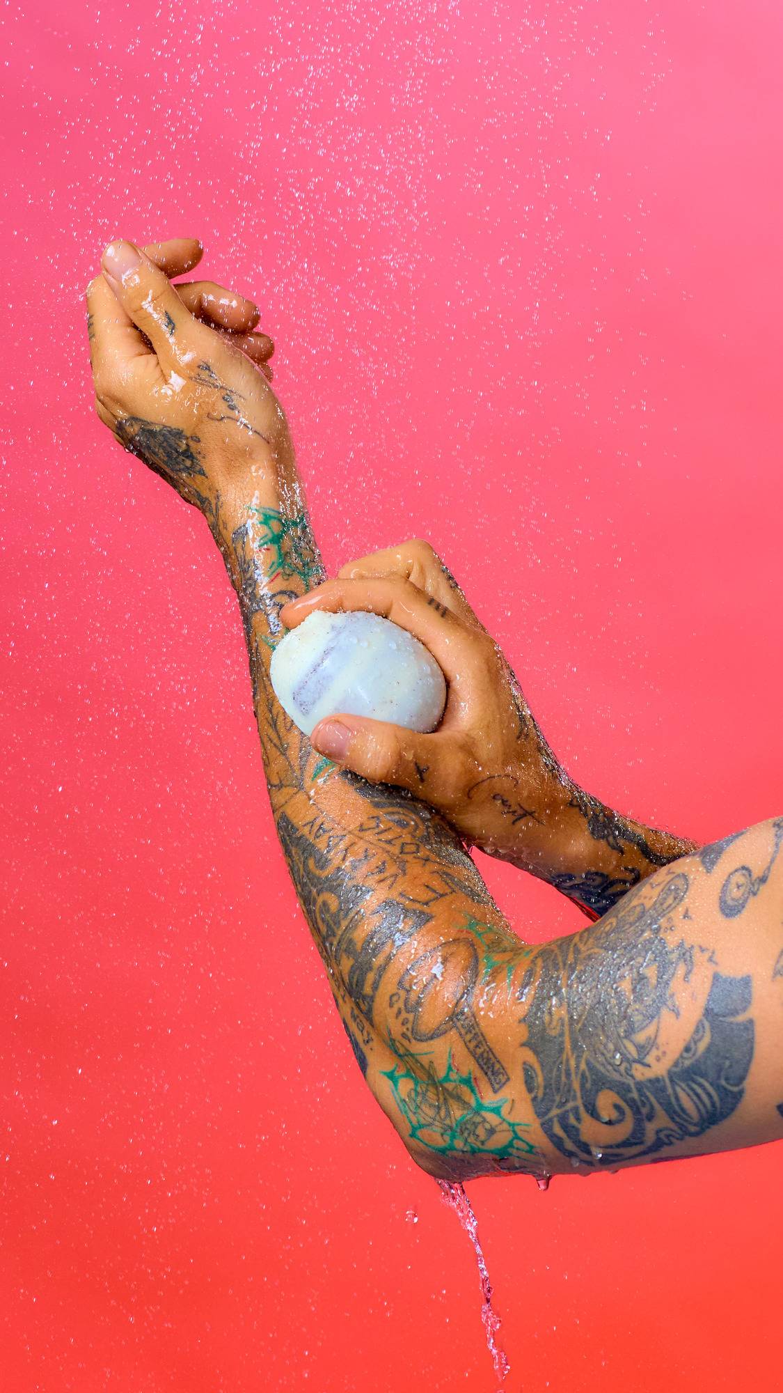 Tattooed arms and hands hold a smooth, light-coloured Scrubee bar under a spray of water against a vibrant pink background. Water droplets cover the glistening skin and stream down from the elbow.