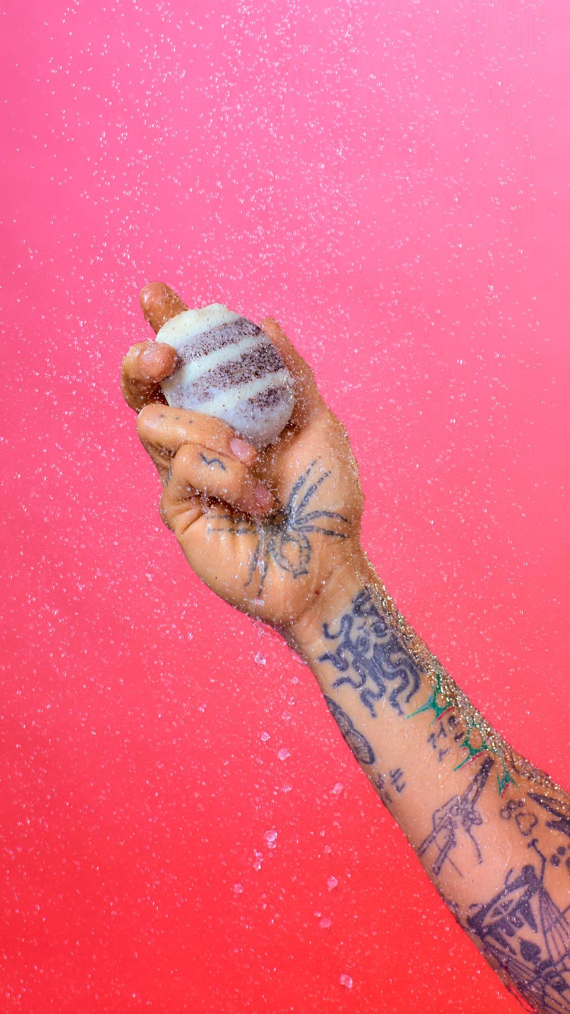 A tattooed arm and hand hold a striped, bee-shaped solid body butter against a bright pink background. Water sprays over the arm, creating a sparkling shower of droplets around the product.