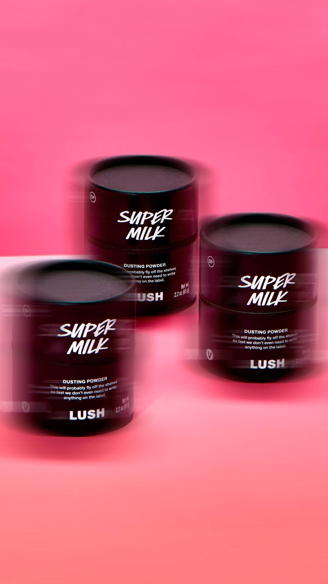 Three black Lush card pots of 'Super Milk' Dusting Powder are arranged with a dynamic horizontal motion-blur effect. They feature bold white text on the front and are set against a vibrant, solid pink background.