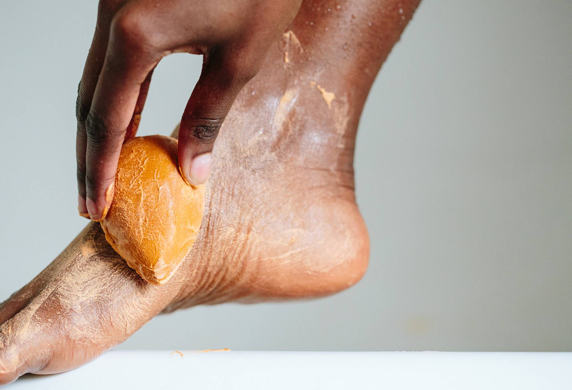 Pumice Power - an orange, teardrop shaped soap - is being rubbed onto the side of a foot resting on the side of a bath.