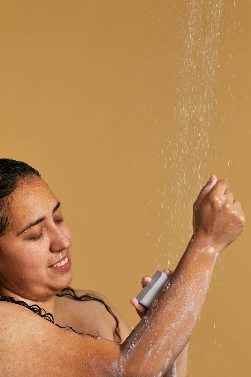 The image shows the model on a sandy, golden background as they are standing under running shower water. The model uses the Sleepy soap slice on their forearm as it begins to lather up with soapy suds.