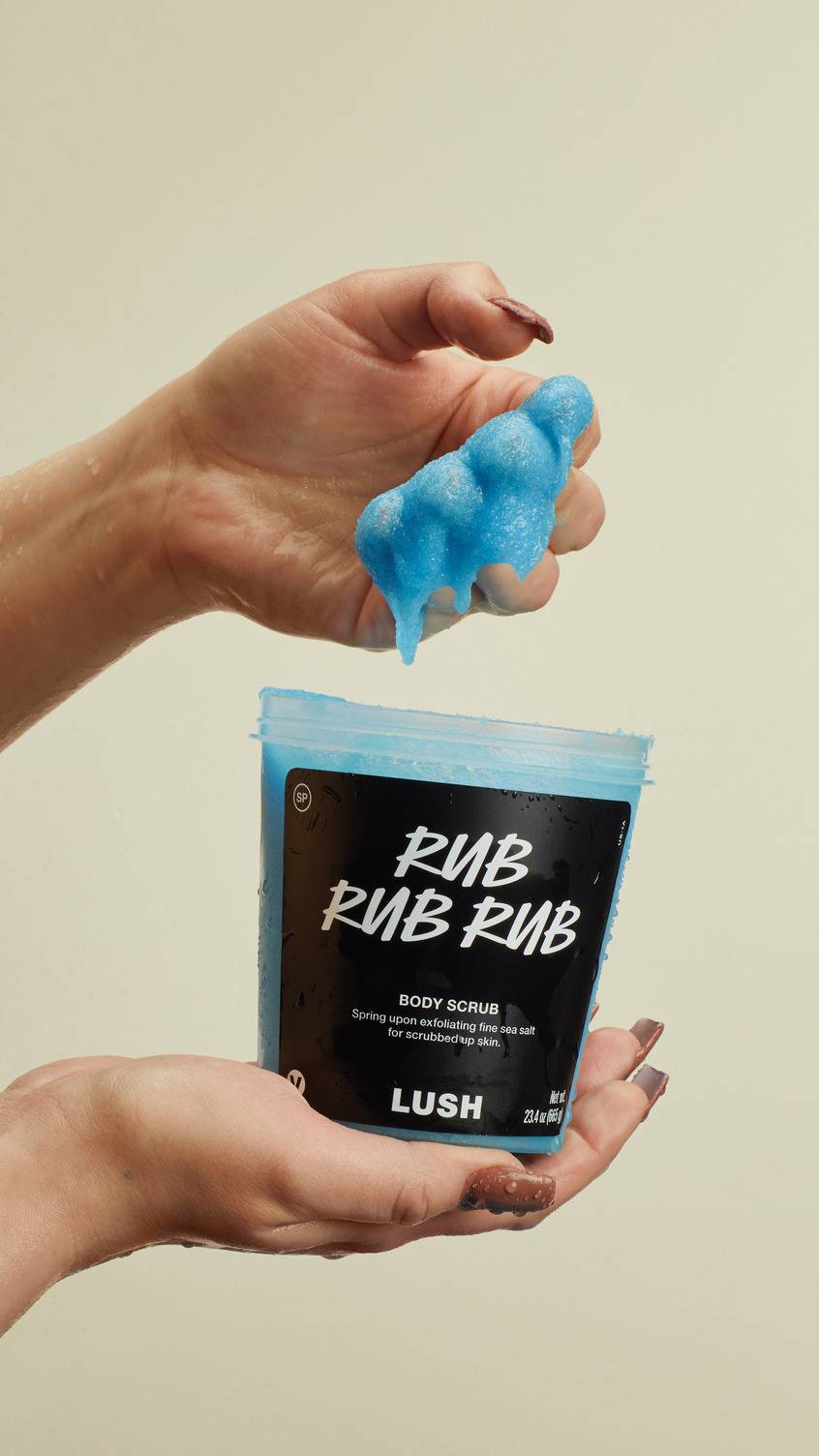 The image shows a close-up of a model's hands holding a clear pot of vibrant blue "Rub Rub Rub" body scrub by Lush. One hand scoops out a thick, textured dollop of the grainy blue scrub, which drips slightly.
