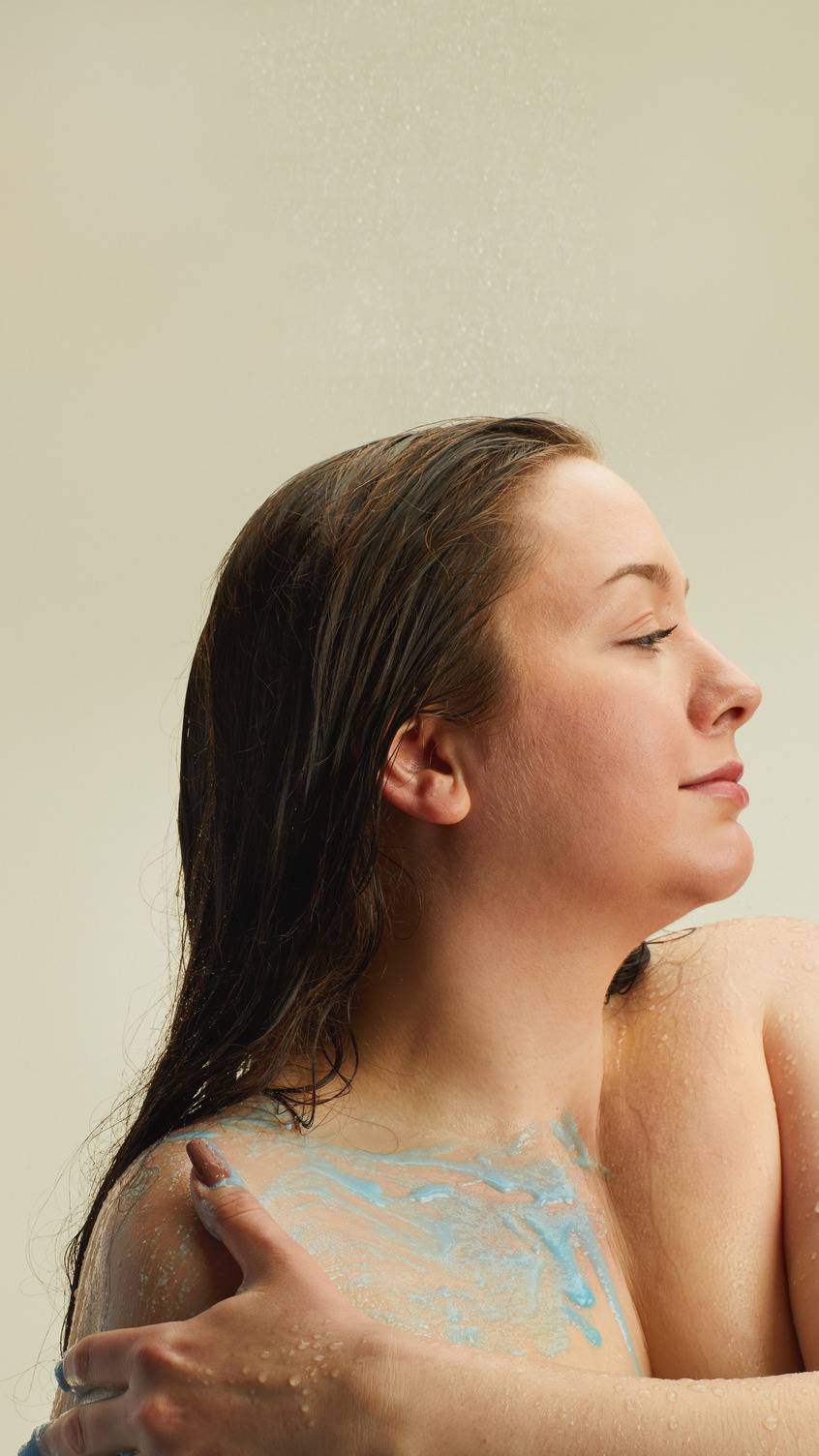 A model with damp hair and eyes closed stands in a shower, applying a vibrant blue body scrub to their arm and shoulder. The textured blue scrub is smeared across their skin under a light spray of water, on a beige background.