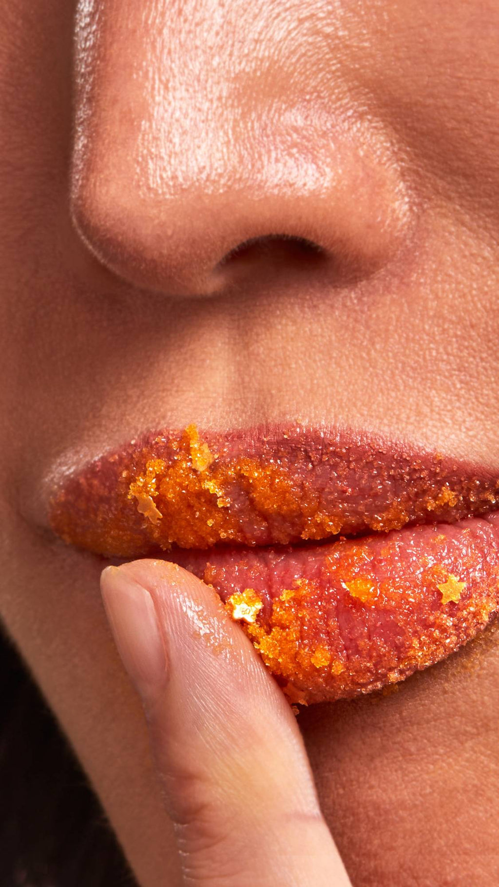 A close-up captures a finger gently applying a granular, bright orange "Sticky Dates" Lip Scrub to a person's lips. The scrub appears sugary and textured, featuring small, star-shaped glitter or confetti pieces embedded within the mixture.