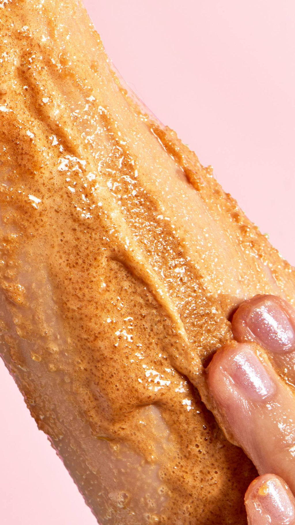 A close-up captures a hand with glittery nails massaging a thick layer of golden-brown "Sticky Dates" Sugar Scrub onto skin. The granular texture of the scrub is visible as it spreads over the wet surface against a pink background.