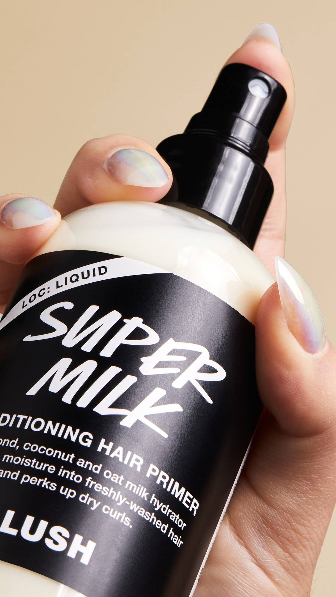 A hand with iridescent manicured nails grips a bottle of "Super Milk" conditioning hair primer. The clear bottle reveals a creamy white liquid inside, while the black label describes the product as an "almond, coconut and oat milk hydrator".