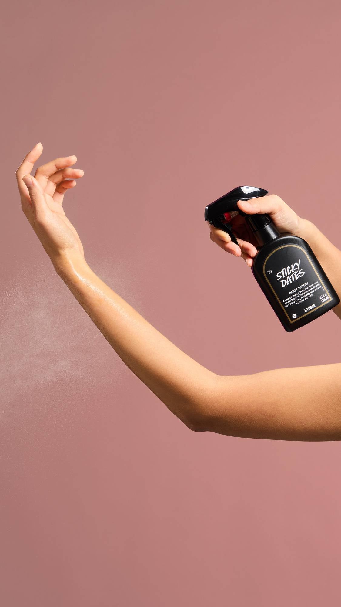 A person sprays a fine mist of "Sticky Dates" Body Spray onto their outstretched forearm against a dusty pink background. The black spray bottle features a label describing the scent as a blend of "benzoin resinoid and sandalwood".