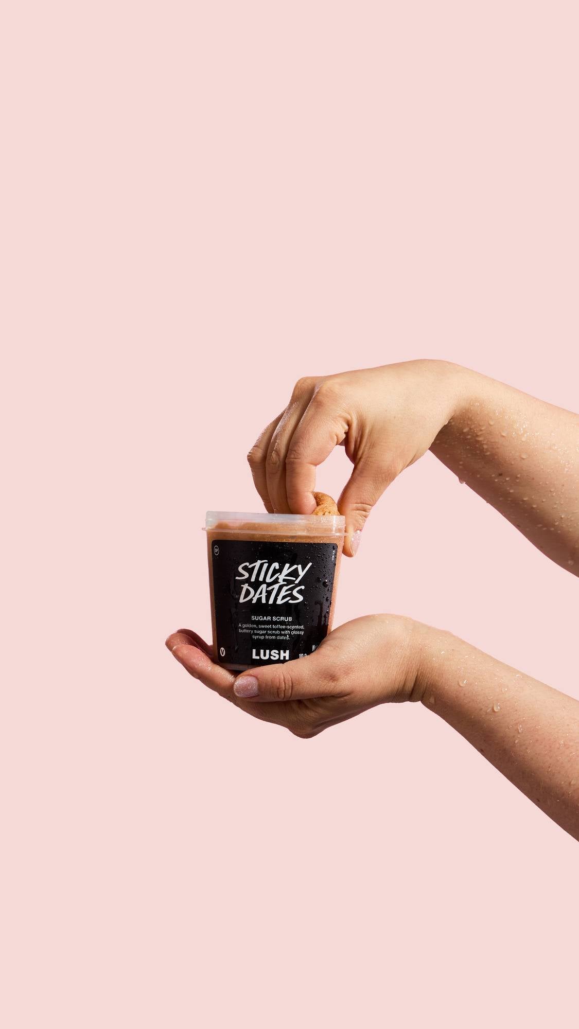Wet hands hold a tub of "Sticky Dates" Sugar Scrub against a pink background. Fingers dip into the open pot, which the label describes as a "sweet toffee-scented, buttery sugar scrub".