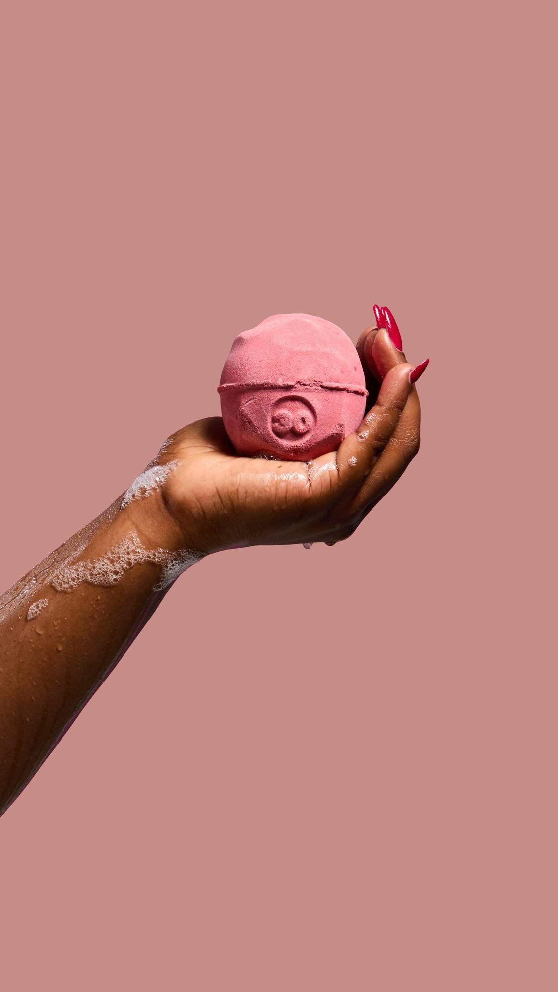 A hand with long red nails and soapy skin holds a pink, spherical "Sticky Dates" bath bomb with a circular indentation against a pink background.