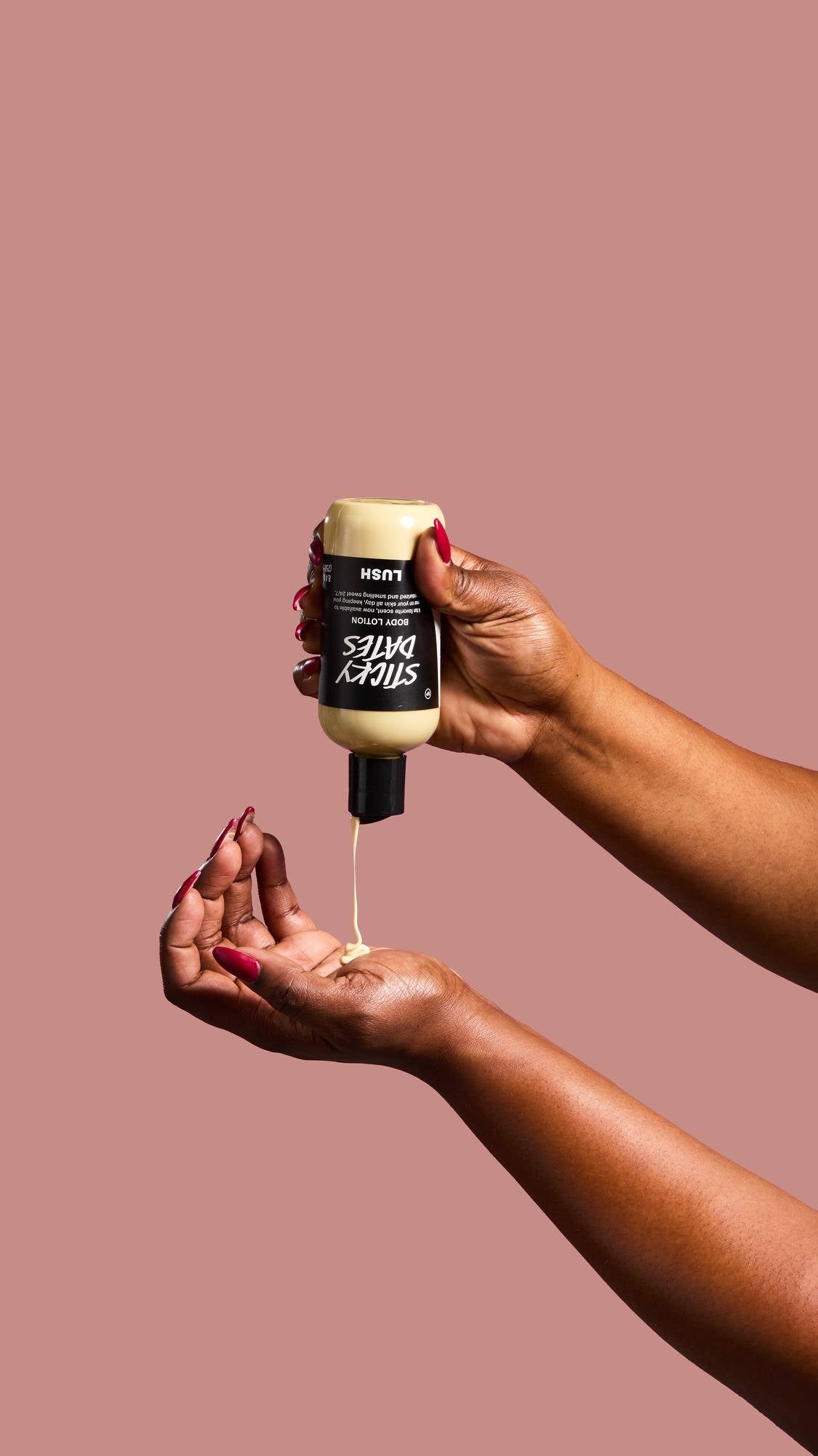 A hand with long red nails holds a bottle of "Sticky Dates" Body Lotion upside down, pouring a stream of creamy, pale yellow liquid into a cupped palm against a dusty pink background.