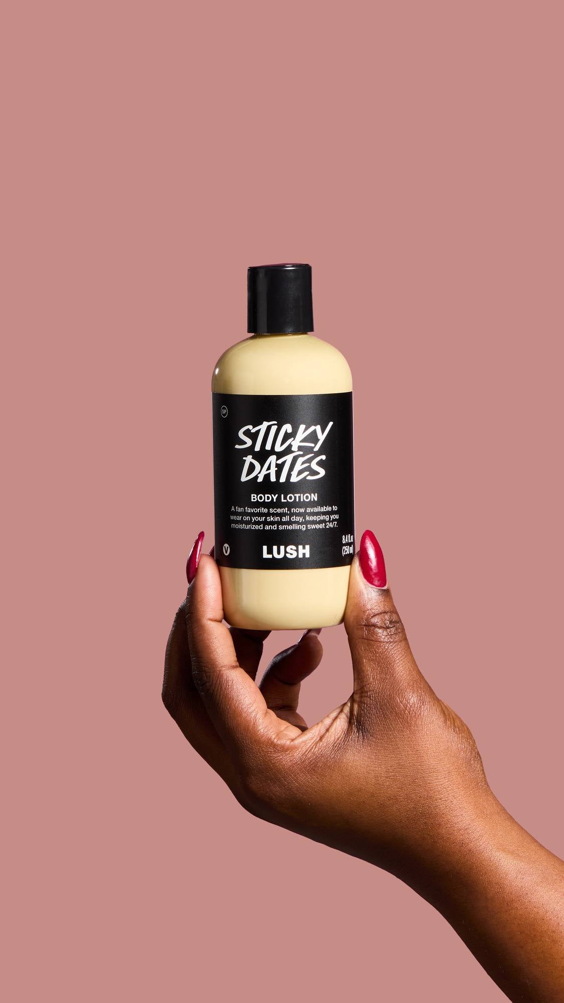 A hand with deep red manicured nails holds a bottle of "Sticky Dates" Body Lotion against a dusty pink background. The black label describes the product as a "fan favorite scent" designed to keep skin "moisturized and smelling sweet 24/7".