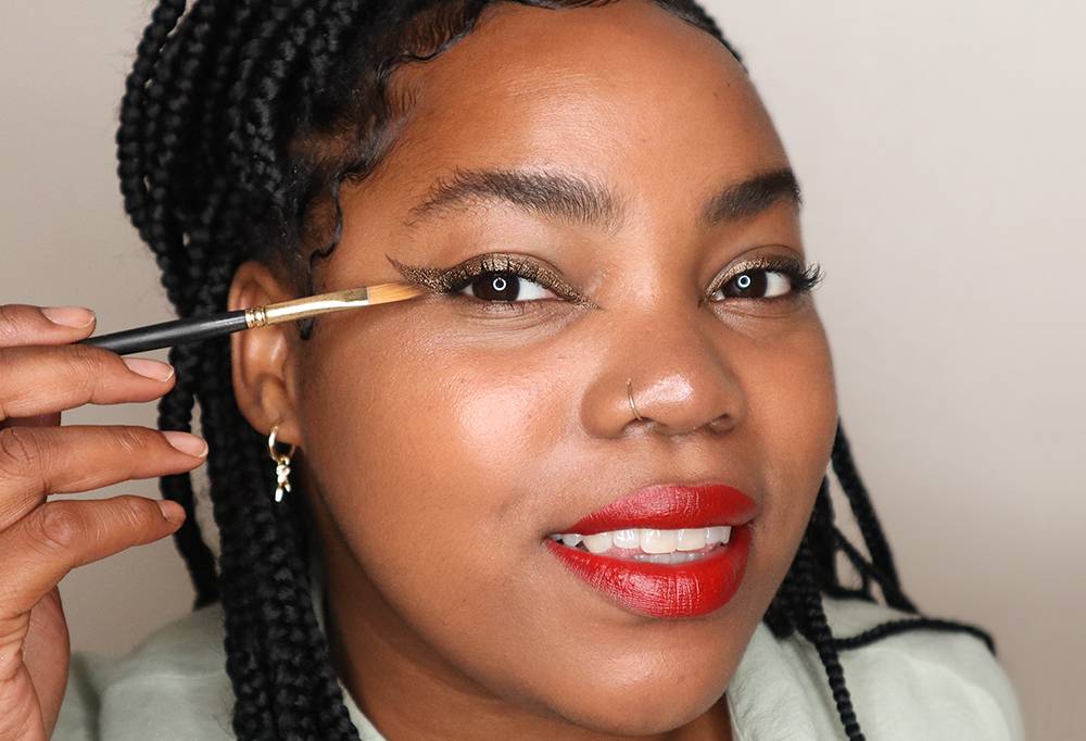 A person smiles as they perfect their winged eyeliner, using Grounded, a bronze eyeliner, and Wing It, an angled liner brush.