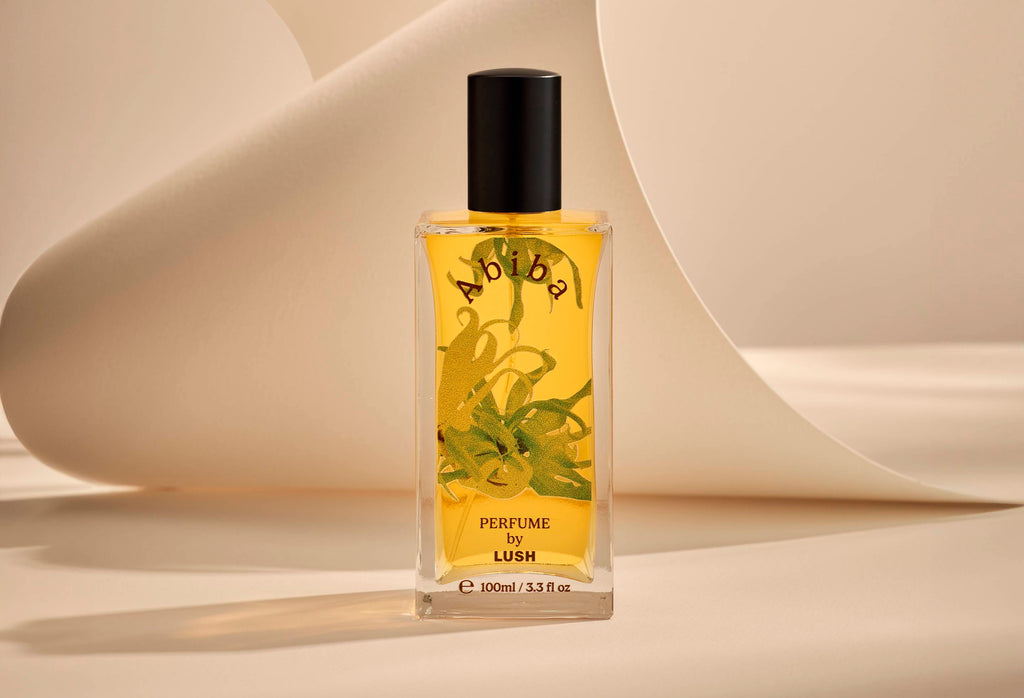 The image shows the Abiba glass perfume bottle. It is sitting on a soft, beige background with curved, cream paper creating soft, gliding shapes behind the product.