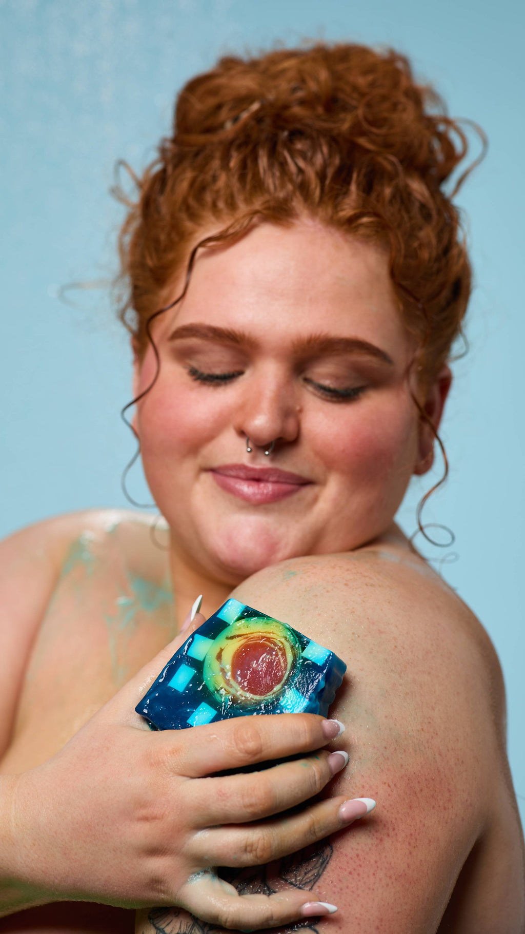 The model is standing under running shower water on a pale blue background. Their curly hair is tied up as they lather their shoulder with the Alban Arthan soap.