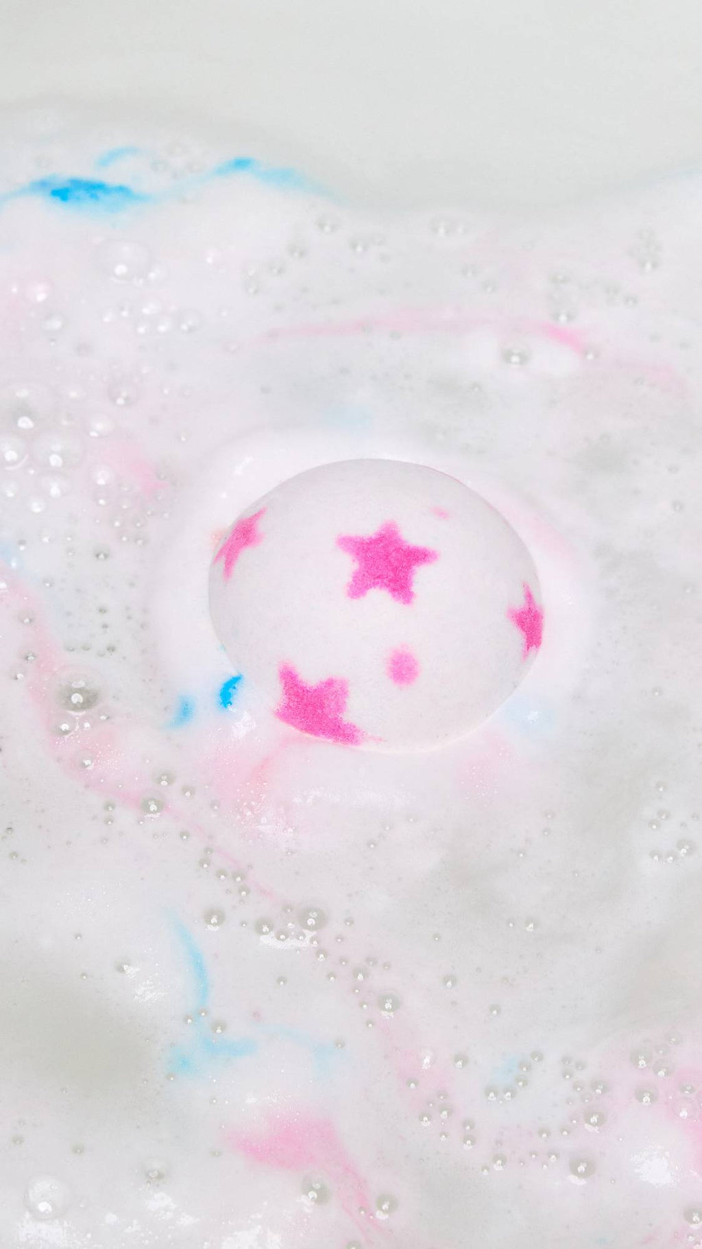 The American Dream bath bomb has just been gently placed into the bath water and is dispersing thick, foamy waves of white with flecks of pink and blue.