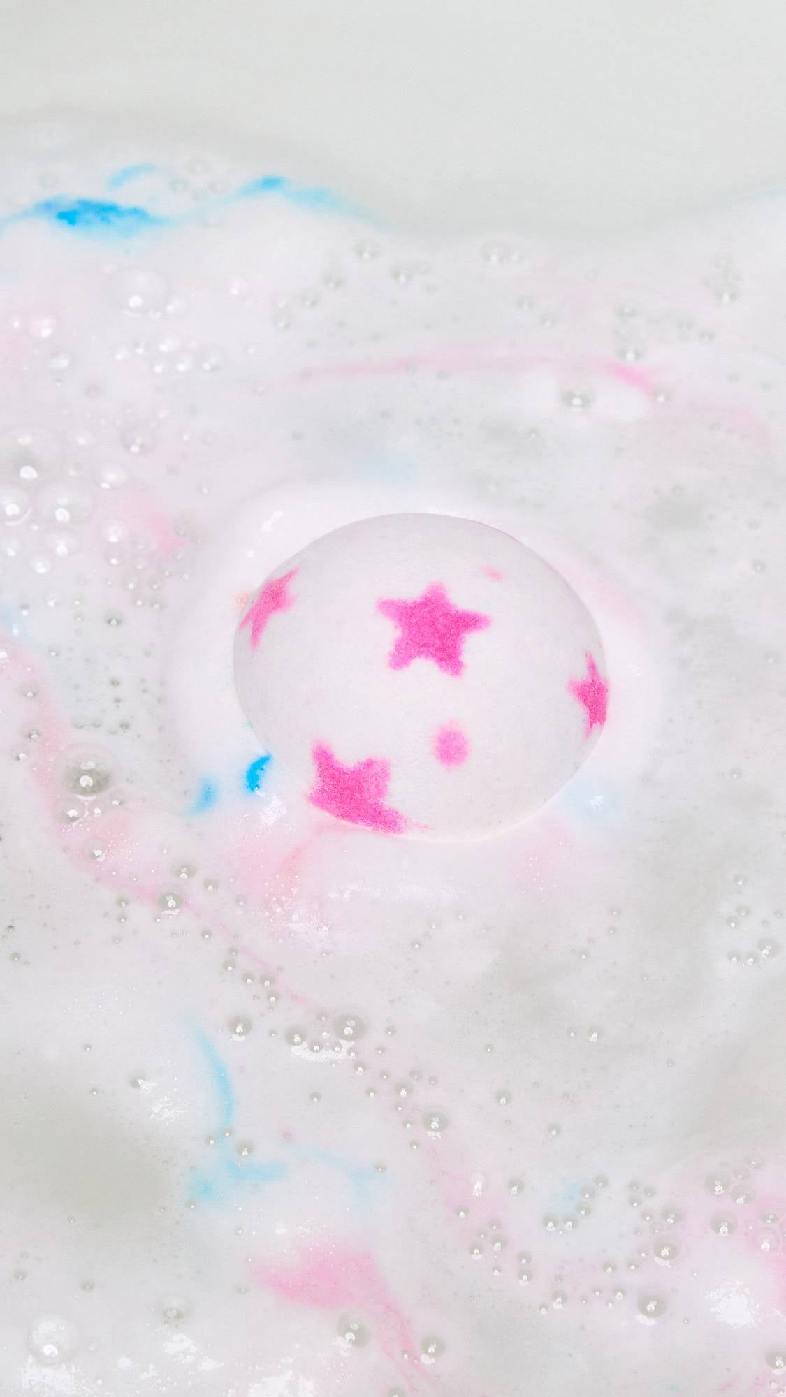 The American Dream bath bomb has just been gently placed into the bath water and is dispersing thick, foamy waves of white with flecks of pink and blue.