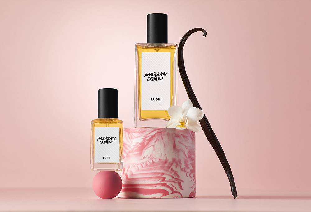 Two perfume bottles balance on a pink ball an cylindrical podium. Next to it is a pink and white flower and a vanilla pod.
