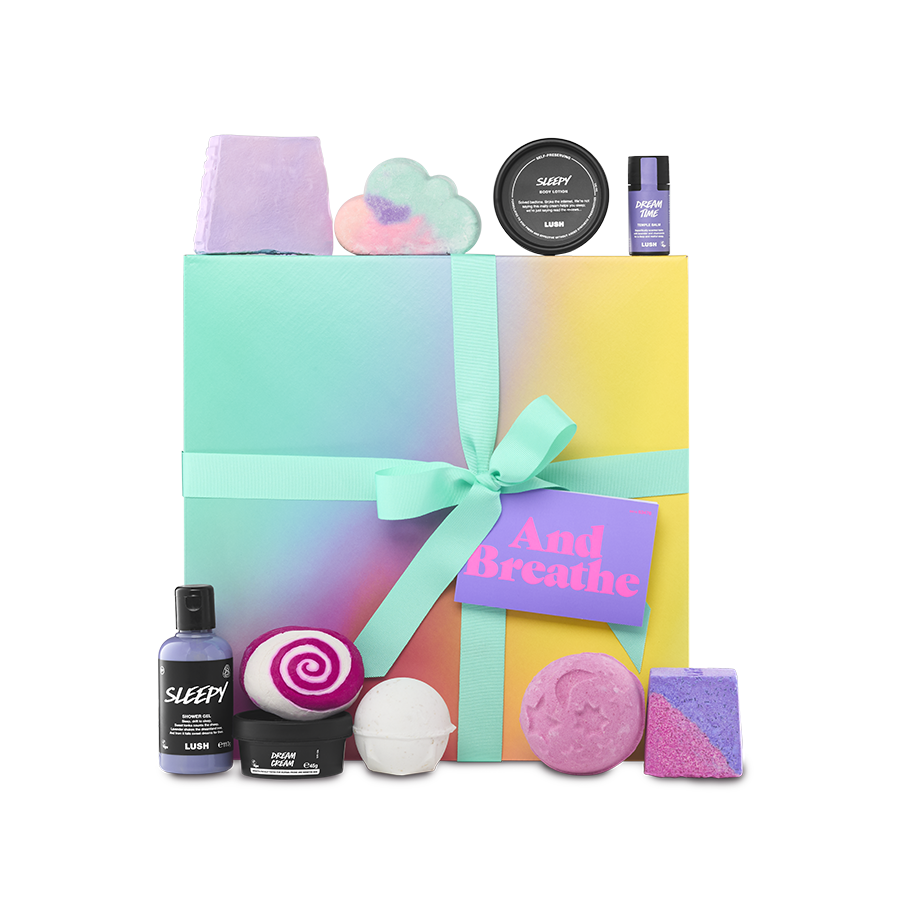 And Breathe. A large, square gift box wrapped in a delicately decorated colour gradient of teal, yellow, pink and orange, tied with a teal ribbon and gift tag. There are ten included Lush products surrounding the gift box.