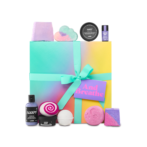 And Breathe. A large, square gift box wrapped in a delicately decorated colour gradient of teal, yellow, pink and orange, tied with a teal ribbon and gift tag. There are ten included Lush products surrounding the gift box.