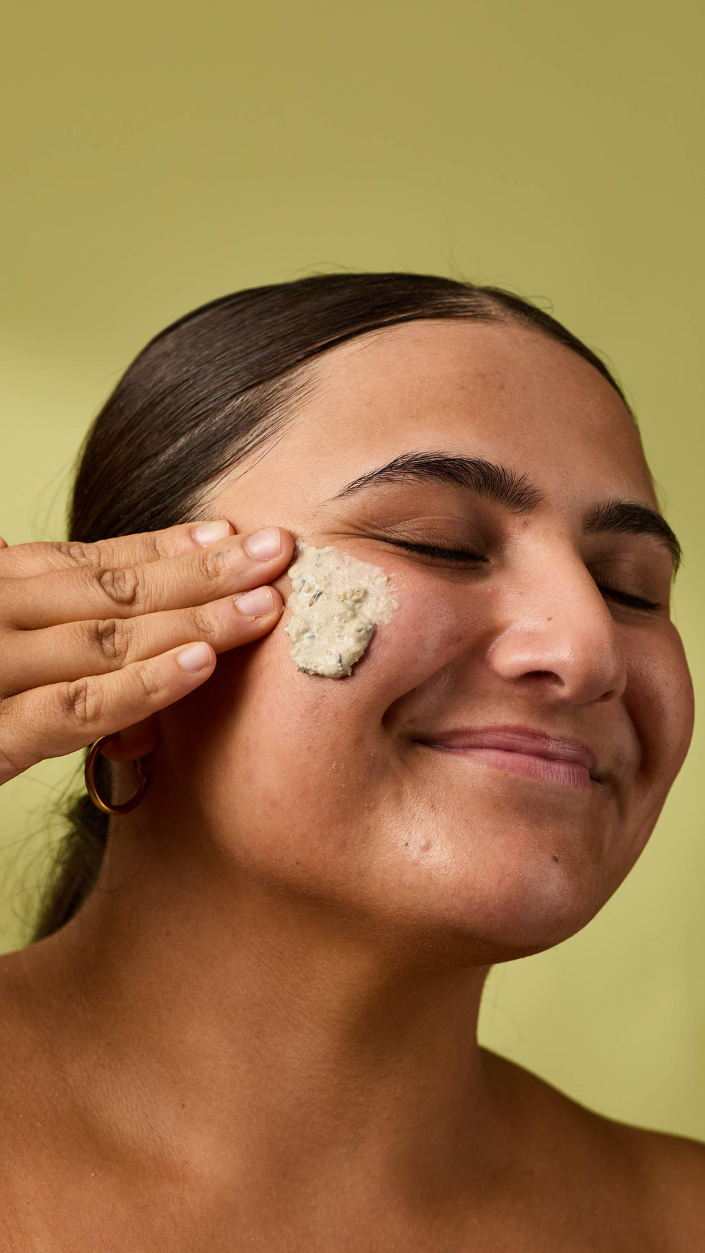 A close-up portrait of a smiling person with eyes closed, gently applying a textured, pale "Angels On Bare Skin" Lush face mask to their cheek with their fingers. Set against a solid, light olive-green background.