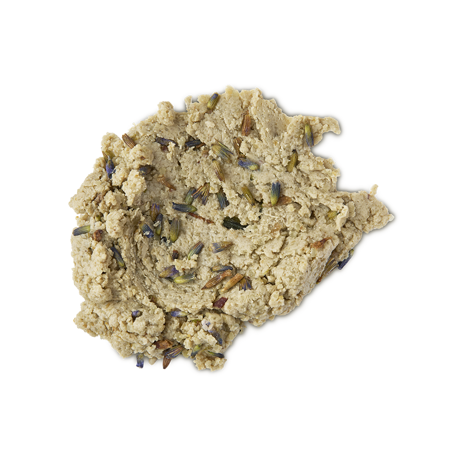 A close-up, top-down studio shot of a light tan, crumbly, dough-like facial cleanser (Lush's Angels on Bare Skin) spread in a dollop on a neutral background. The textured paste is visibly studded with small, dried purple and brown lavender buds