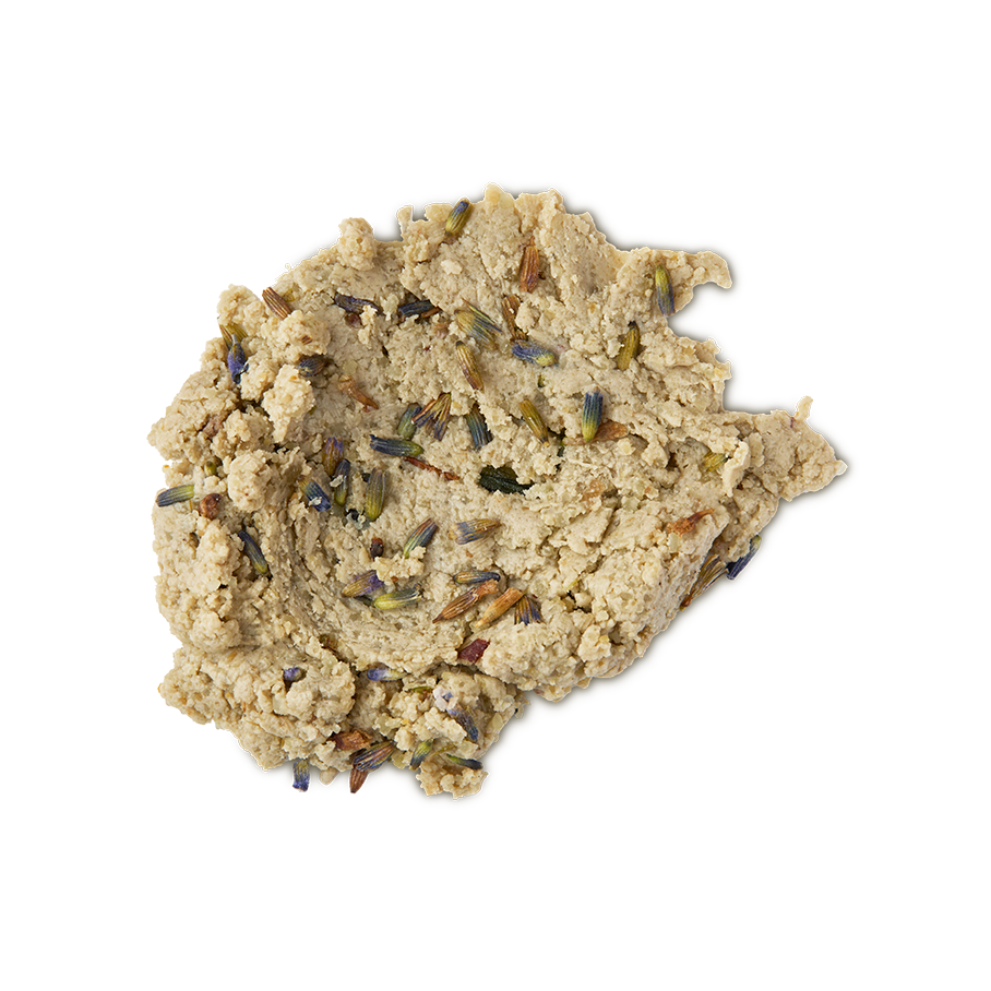 A close-up, top-down studio shot of a light tan, crumbly, dough-like facial cleanser (Lush's Angels on Bare Skin) spread in a dollop on a neutral background. The textured paste is visibly studded with small, dried purple and brown lavender buds