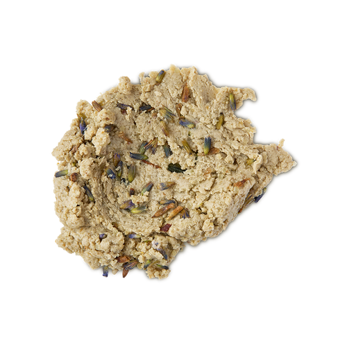 A close-up, top-down studio shot of a light tan, crumbly, dough-like facial cleanser (Lush's Angels on Bare Skin) spread in a dollop on a neutral background. The textured paste is visibly studded with small, dried purple and brown lavender buds
