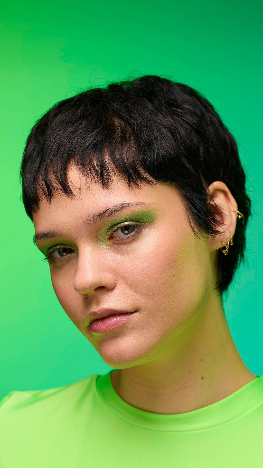 The image shows a close-up of the model's face on a pale blue-to-green gradient background. The model's make-up is done with the focal point being a subtle, jade-green, shimmering eyeshadow made by the Aurora Glitter Crumble.