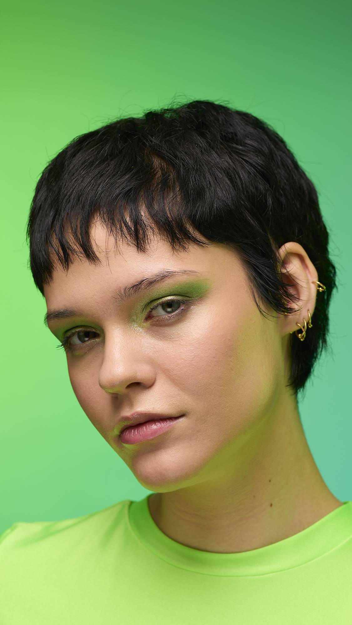 The image shows a close-up of the model's face on a pale blue-to-green gradient background. The model's make-up is done with the focal point being a subtle, jade-green, shimmering eyeshadow made by the Aurora Glitter Crumble.