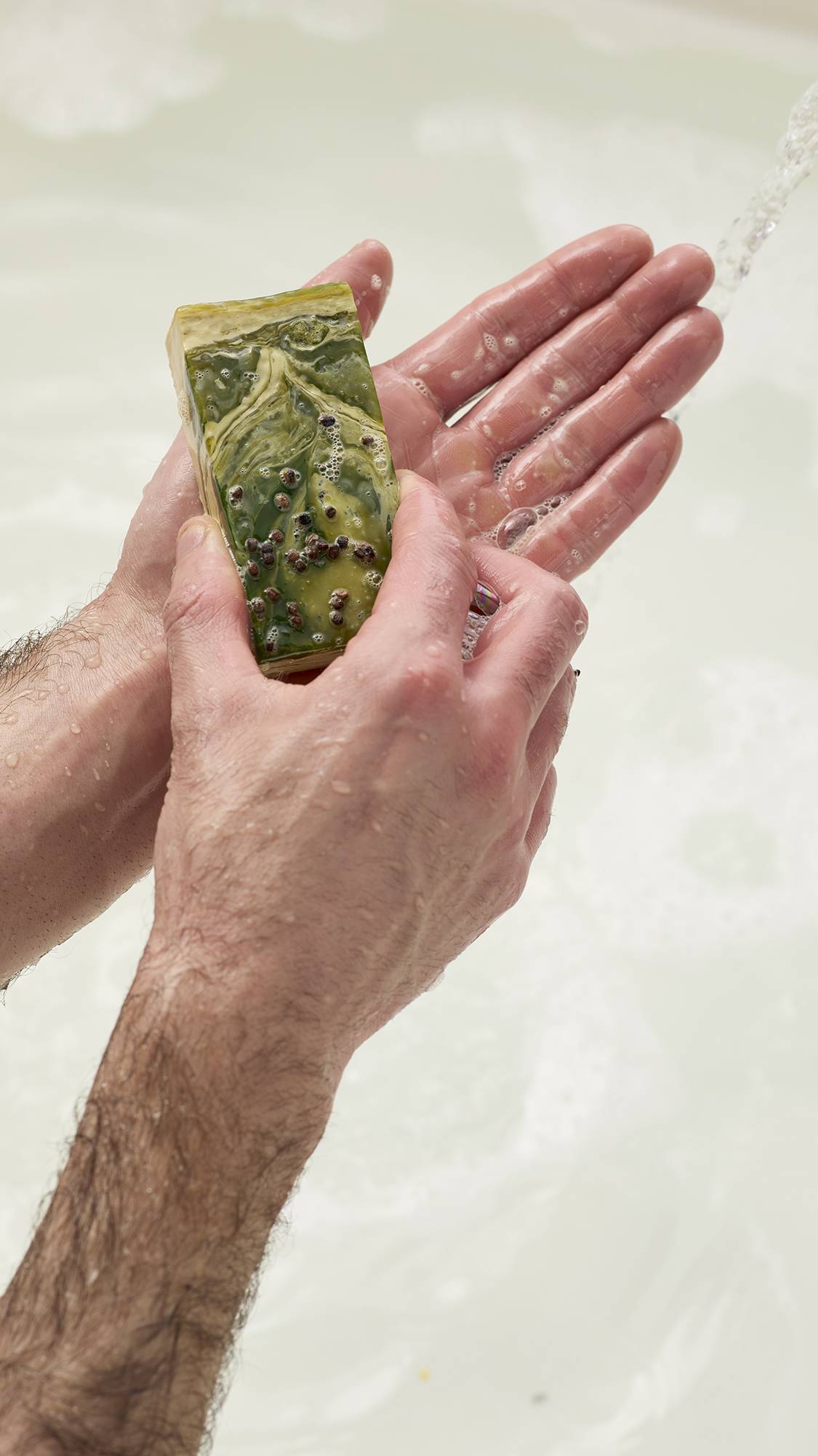 Hands lather the green and gold marbled Baklava soap under a stream of water. Dark seeds are visible on the soap's surface. Set against a soft white background.