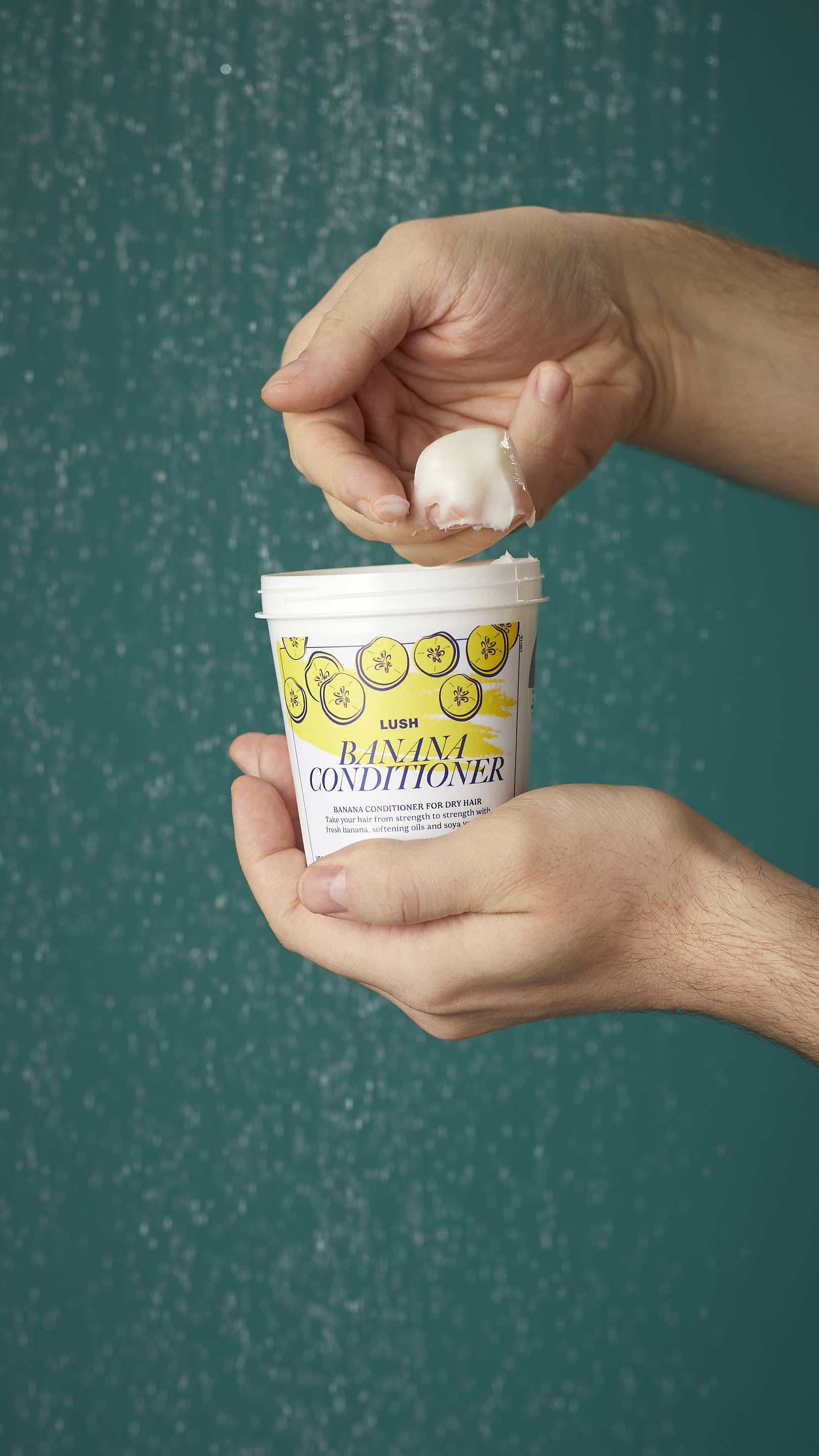 The image shows the model's arms stretched into the centre holding the white, LUSH Version-themed packed pot of the Banana conditioner. One of their hands is scooping the product out. There is a teal background.
