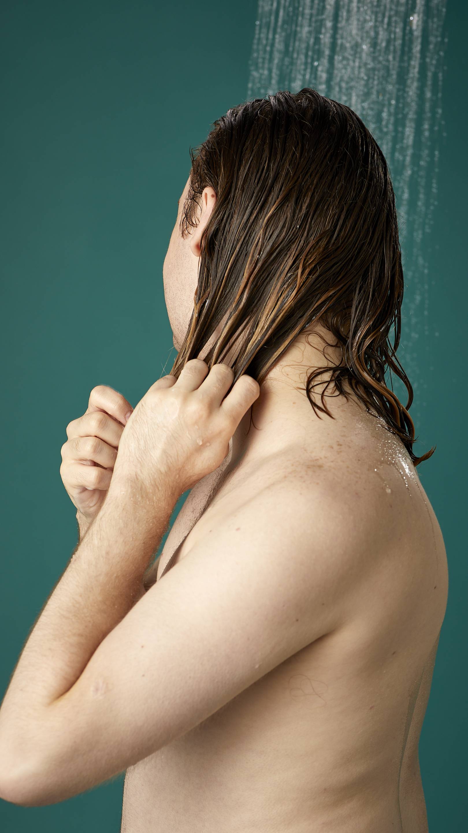 The image shows the model from the side while facing away, under running shower water. Their hair is wet and has been shampooed already. They are now running the Banana conditioner through the lengths and ends of their hair.