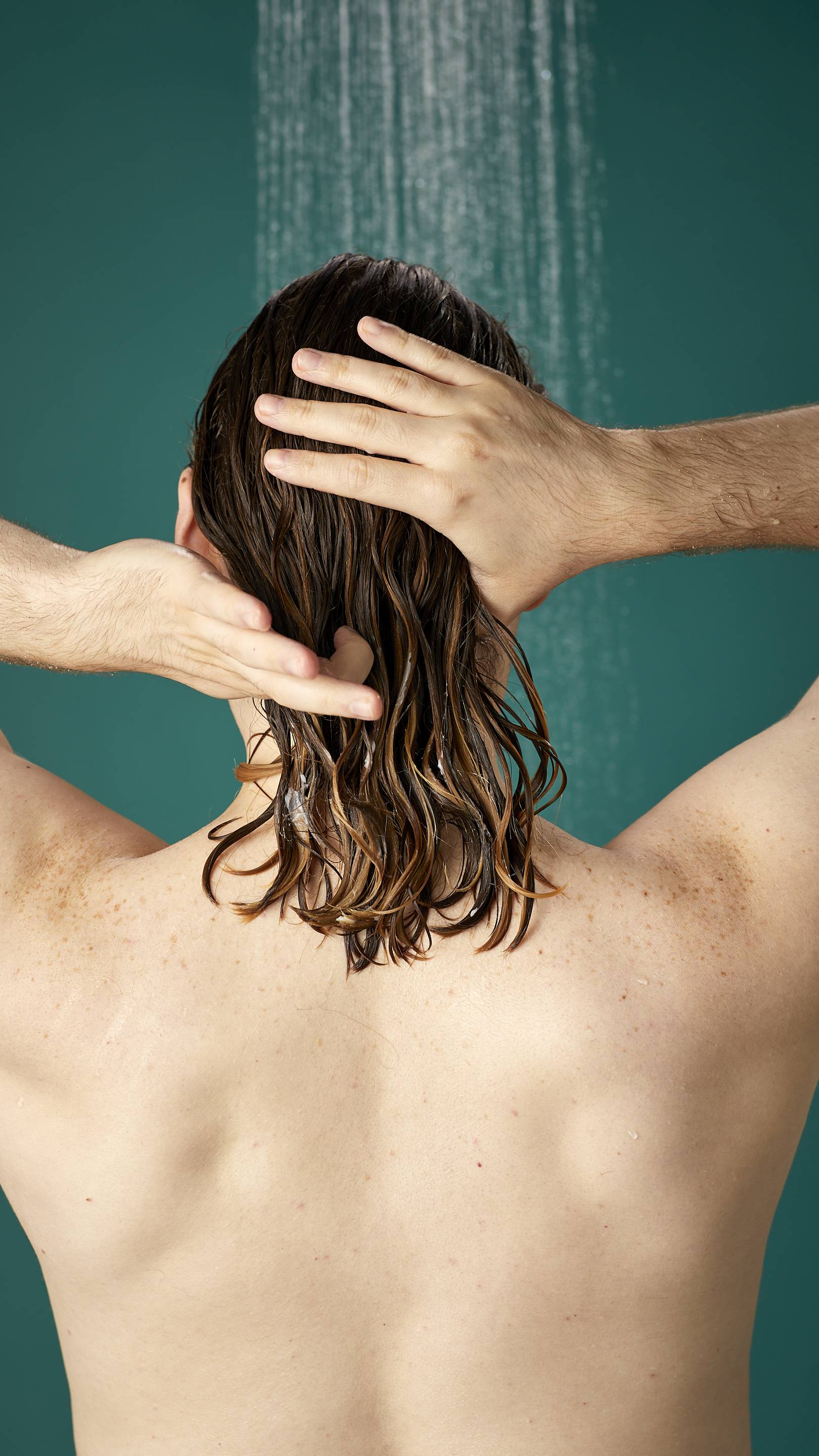 The image shows the model facing away from the camera while standing under running shower water. Their hair is wet and has been shampooed already. They are now running the Banana conditioner through the lengths and ends of their hair.