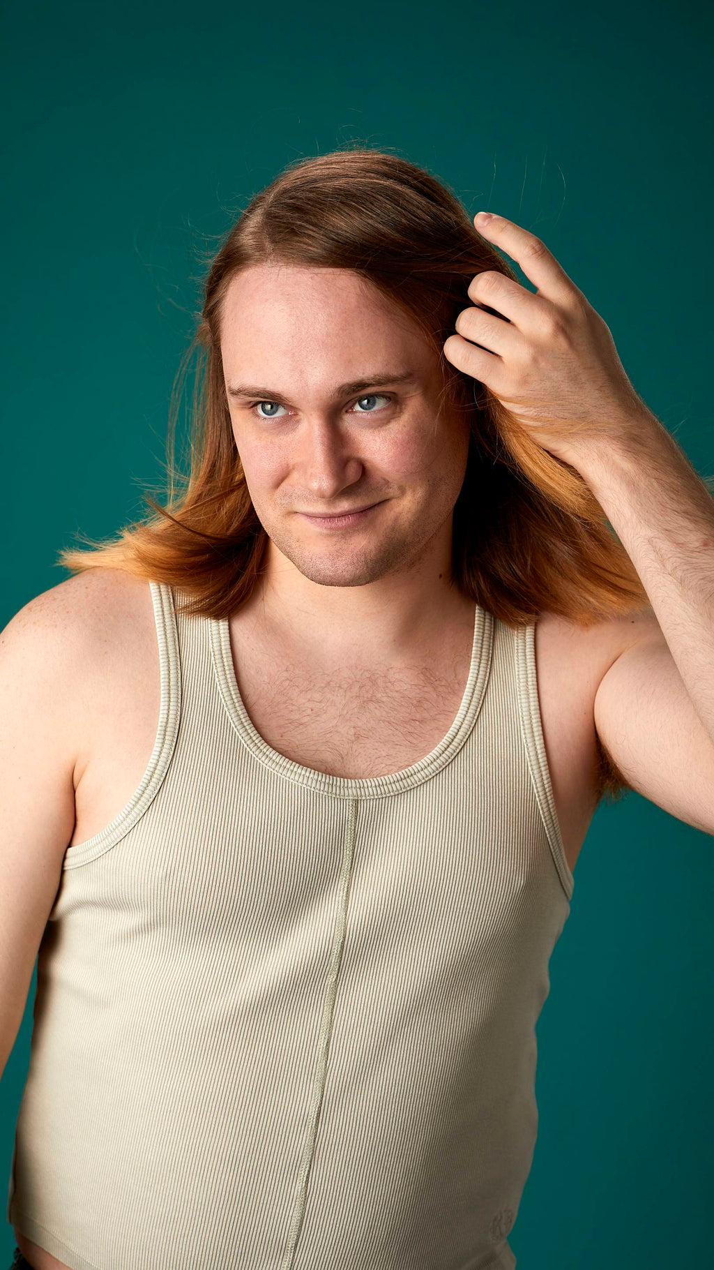 The model is on a teal background with a cream-coloured tank top. They have light brown, shoulder-length, freshly-dried hair which looks shiny and healthy and is being fanned with air.