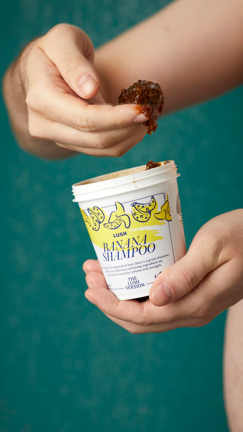 The image shows the model's arms stretched into the centre holding the white, LUSH Version-themed packed pot of the Banana shampoo. One of their hands is scooping the product out. There is a teal background.