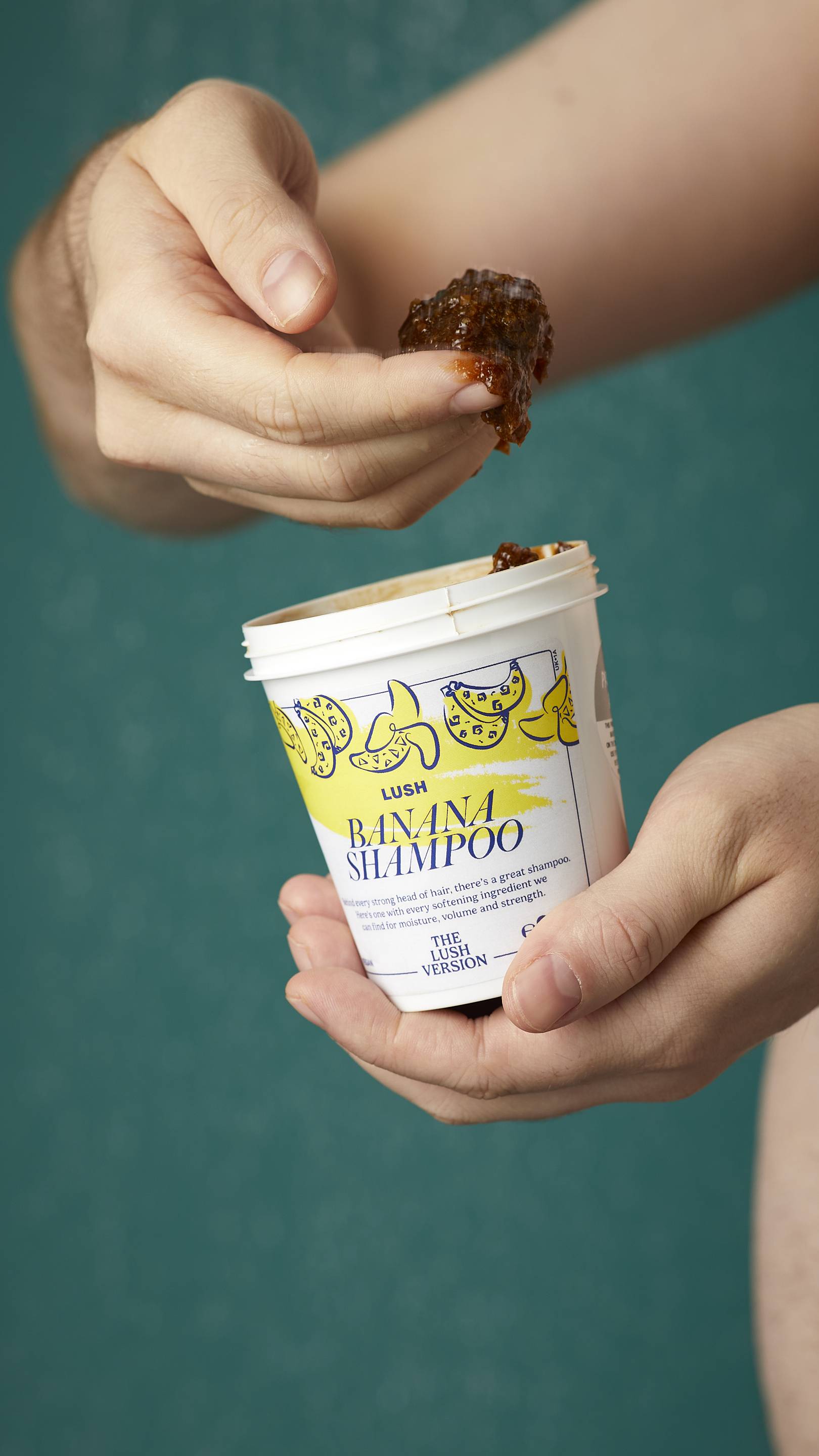 The image shows the model's arms stretched into the centre holding the white, LUSH Version-themed packed pot of the Banana shampoo. One of their hands is scooping the product out. There is a teal background.
