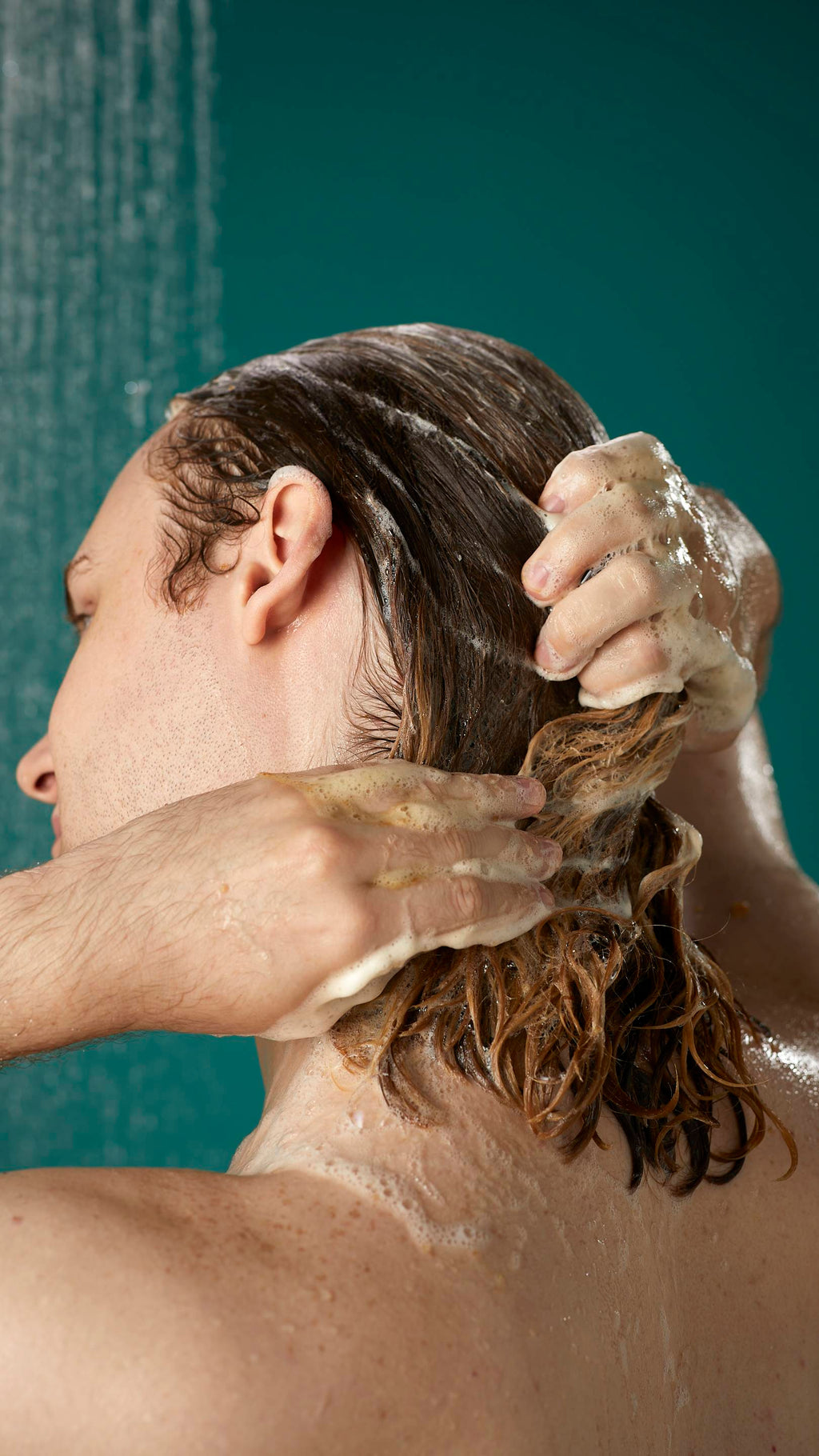 The image shows the rear view of the model's shoulder-length hair which is wet and lathered in suds as they lather the Banana shampoo into their roots. There is a teal background with running shower water.
