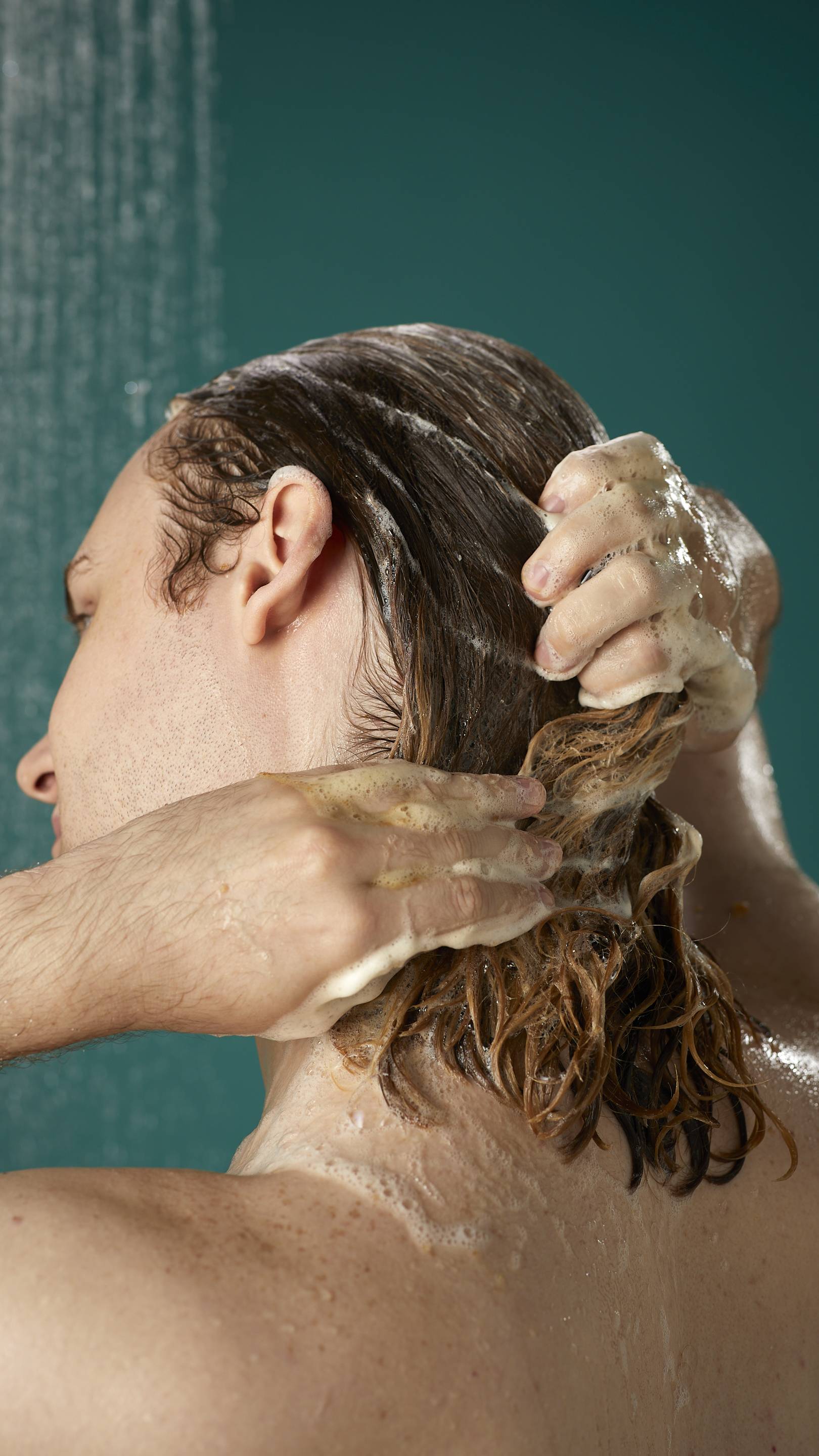 The image shows the rear view of the model's shoulder-length hair which is wet and lathered in suds as they lather the Banana shampoo into their roots. There is a teal background with running shower water.