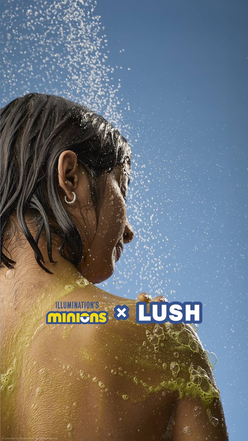 The image shows the model standing under running shower water as they face away from the camera. They have lathered the Bananaaaa shower cream over their shoulder and back. The Minions X LUSH logo is at the bottom.