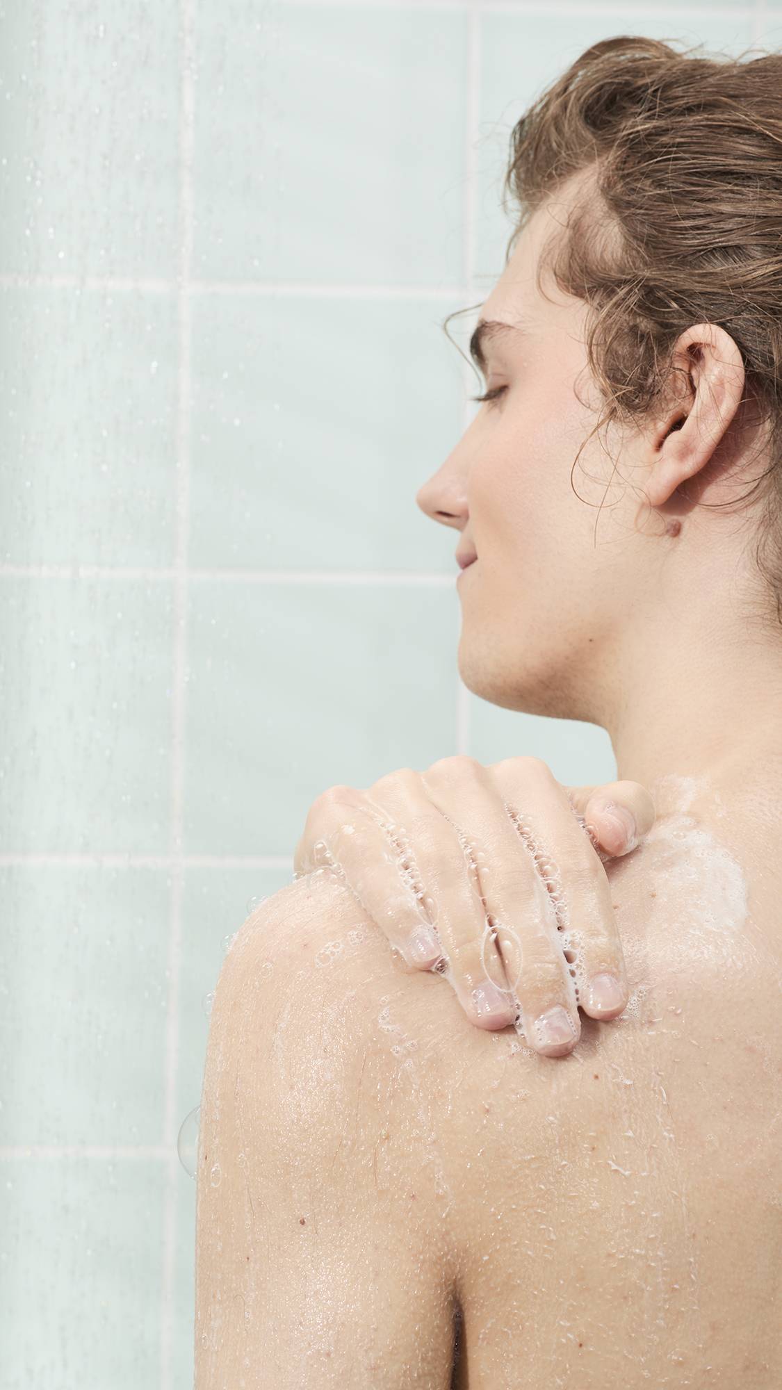 A person's side profile in the shower, applying a light, bubbly soap lather to their shoulder. The background features light mint green wall tiles and the soft spray of falling water.