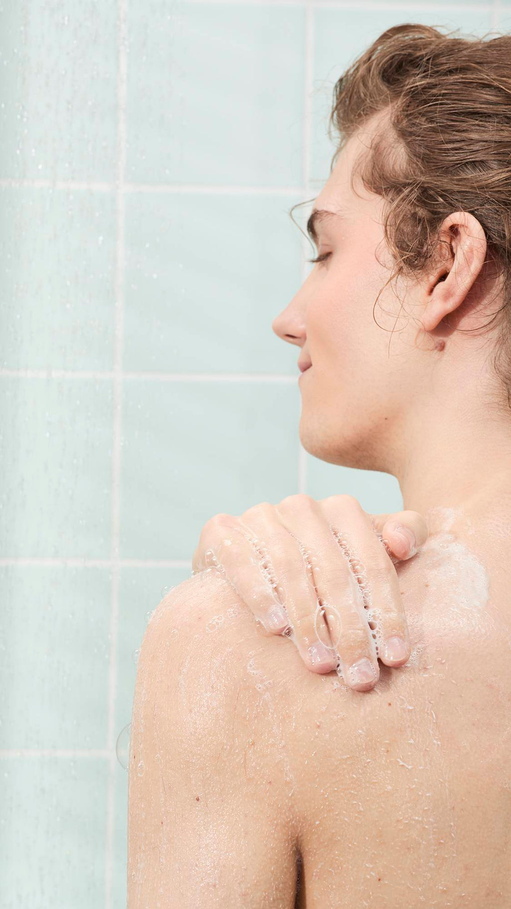 A person's side profile in the shower, applying a light, bubbly soap lather to their shoulder. The background features light mint green wall tiles and the soft spray of falling water.