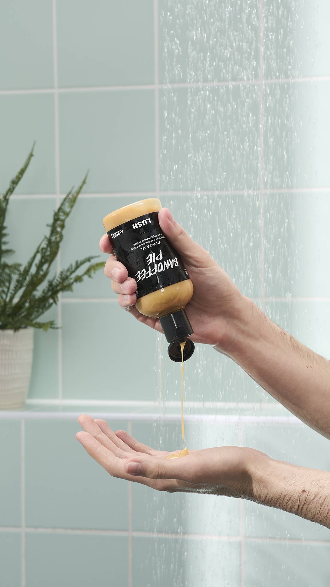 A person's hands are shown in a shower, with one hand holding an upside-down bottle of Lush Banoffee Pie shower gel. A thick stream of the creamy yellow gel is being squeezed into the palm of the other hand against a background of mint green tiles.
