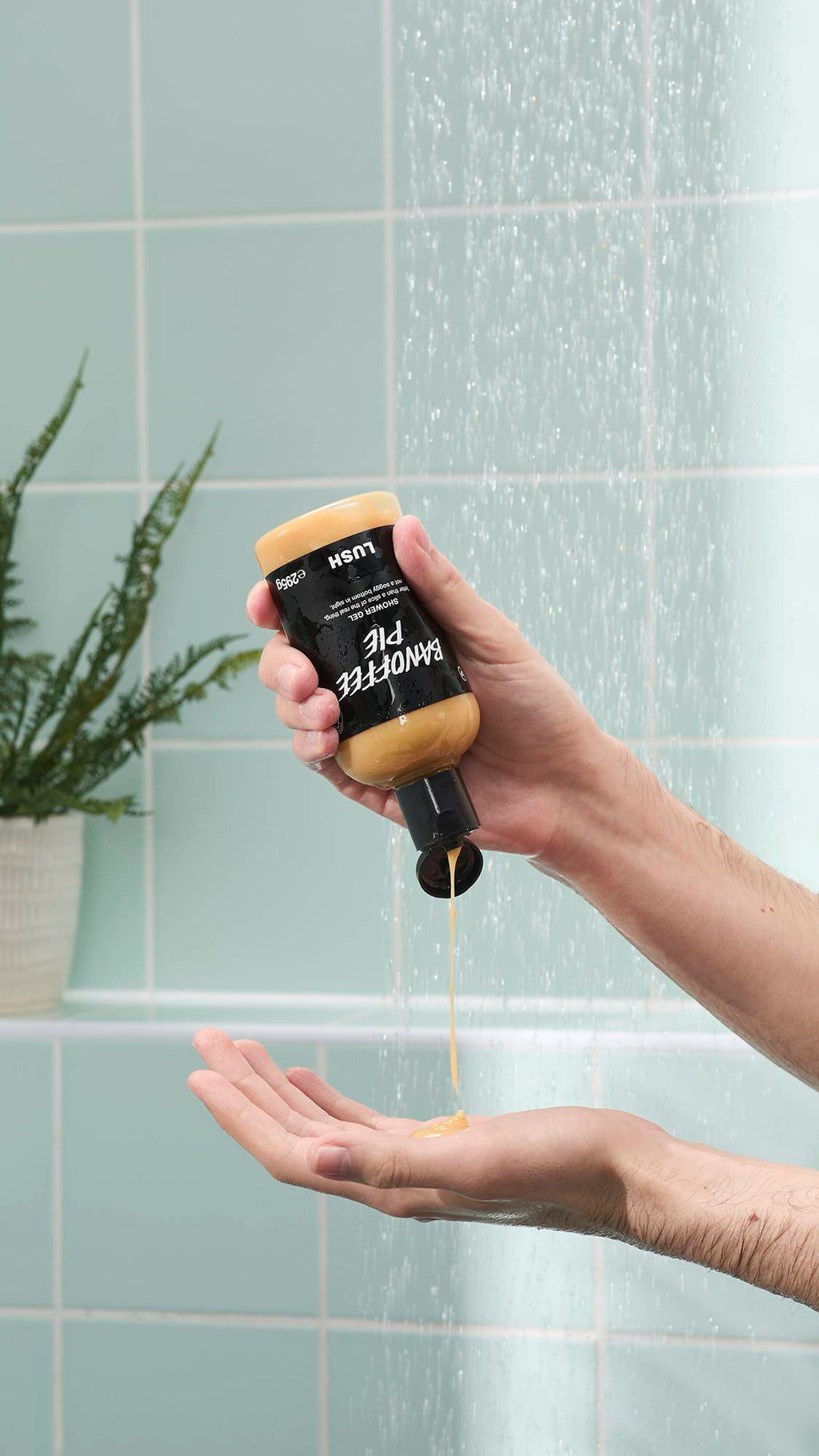 A person's hands are shown in a shower, with one hand holding an upside-down bottle of Lush Banoffee Pie shower gel. A thick stream of the creamy yellow gel is being squeezed into the palm of the other hand against a background of mint green tiles.