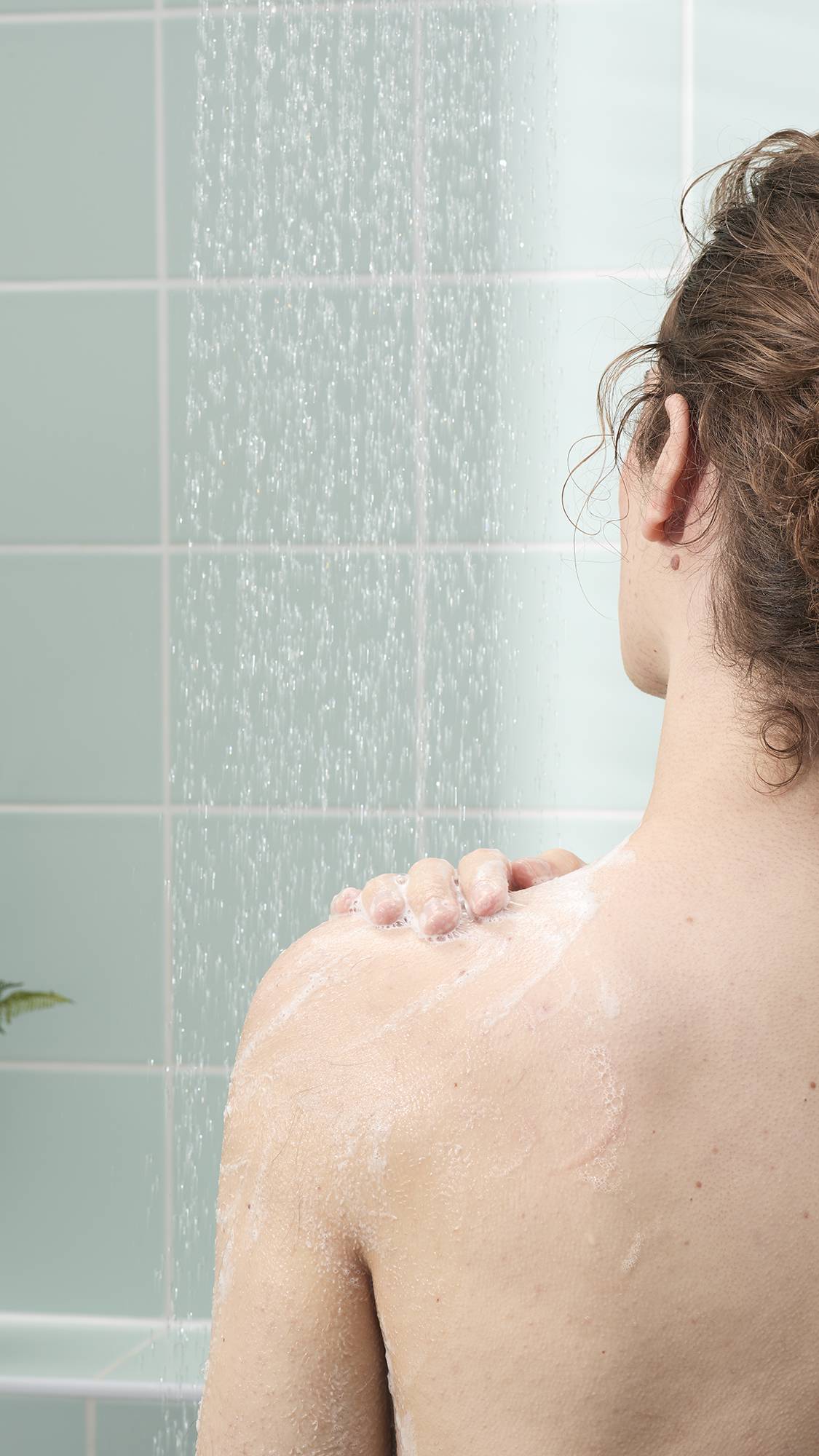 A person's back and shoulder are shown from behind in a shower as they apply a light, creamy soap lather. The background features light mint green wall tiles and the steady, soft spray of falling water.