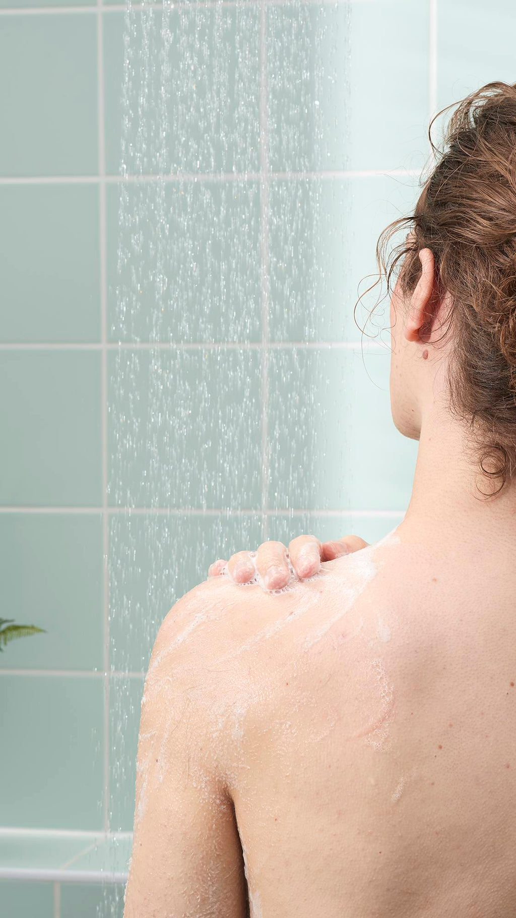 A person's back and shoulder are shown from behind in a shower as they apply a light, creamy soap lather. The background features light mint green wall tiles and the steady, soft spray of falling water.