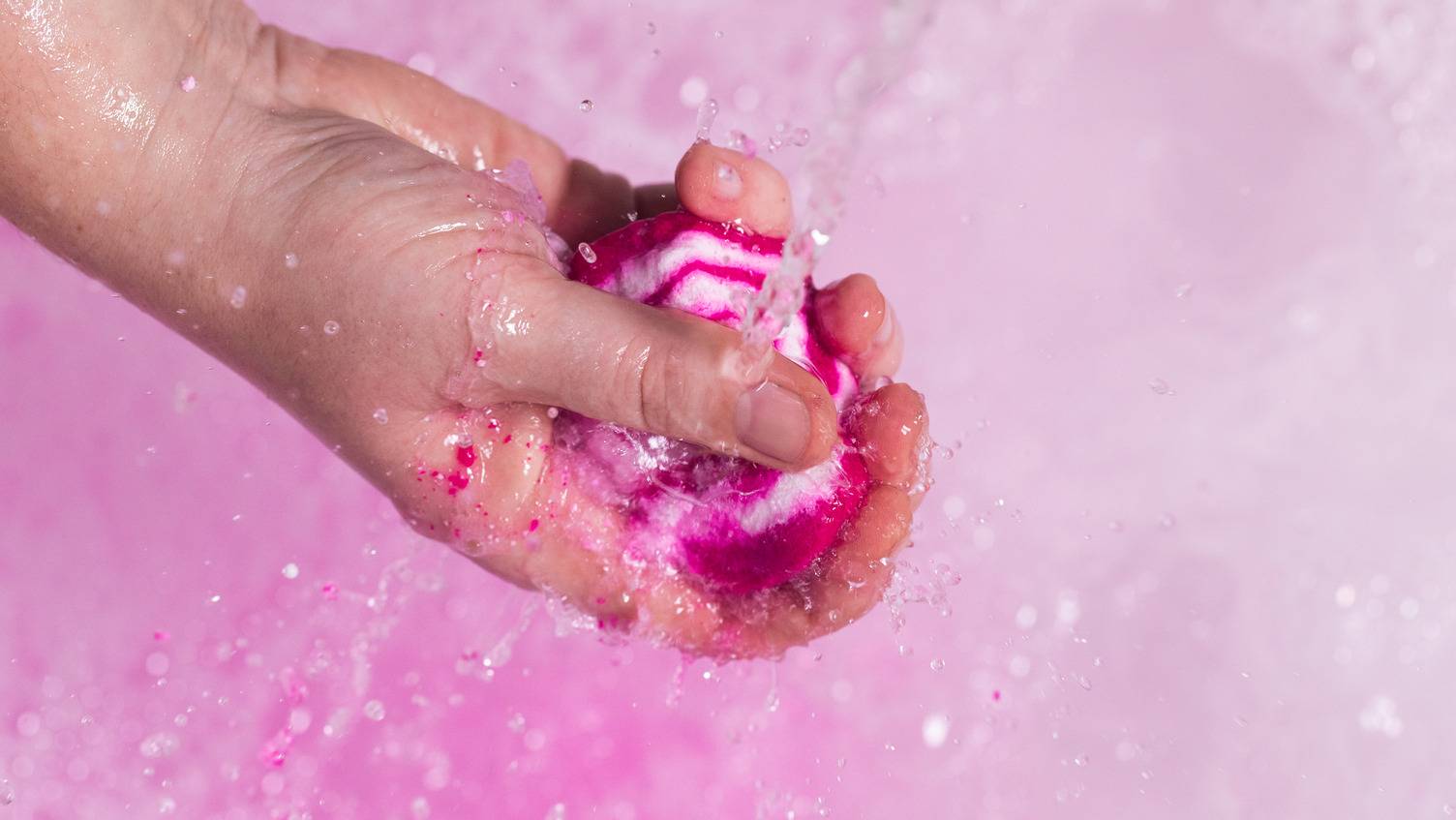 The image shows the model's hand as it crumbles a mini The Comforter bubble bar under running tap water to produce a mountain of bubbles below and vibrant, pink bath water.