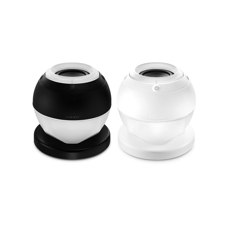 Bath Bot. Two slick, bath bomb-shaped speaker gadgets with a flat base and modern design. One is fully white and the other is black and white. There is an embossed power button visible and the word "LUSH".
