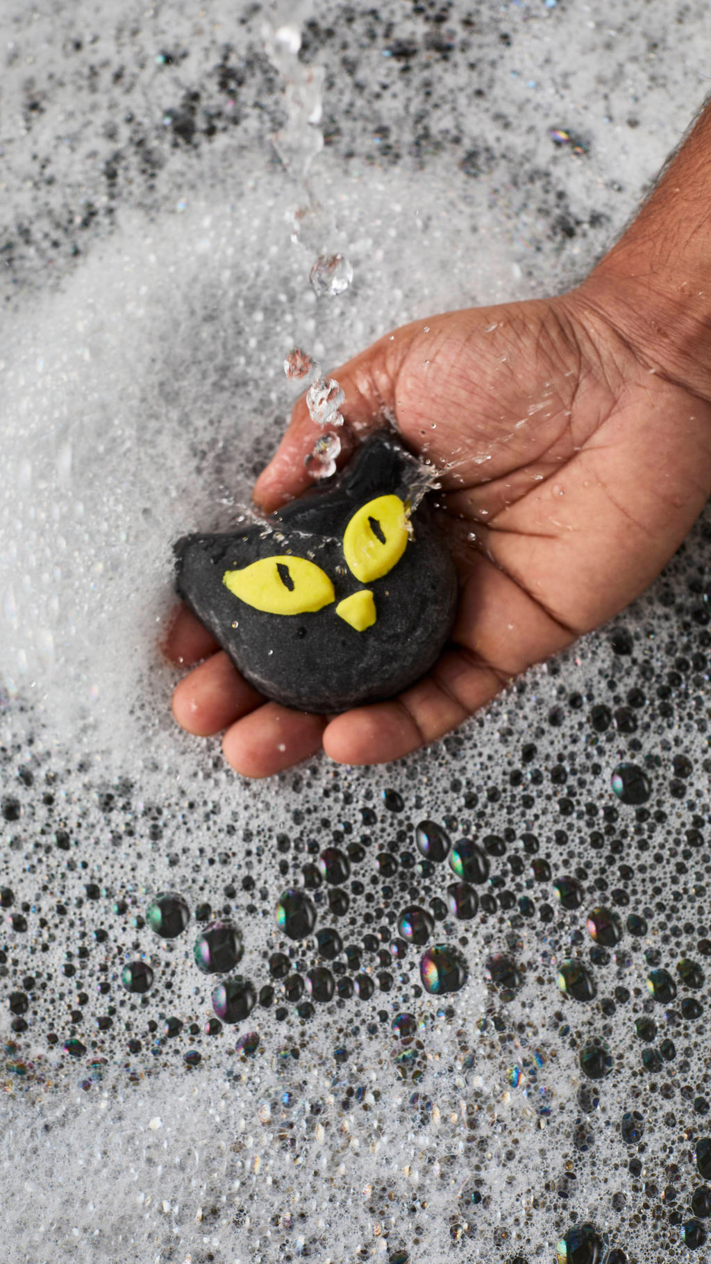 A person's hand holds a black cat-shaped bubble bar with bright yellow eyes under a stream of running tap water. The water splashes off the bar into a bath covered with a thick, frothy layer of white bubbles.